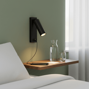 Wall-mounted black reading light with USB port beside a wooden bedside table, featuring a phone and water glasses.
