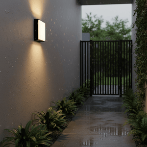 Kwele Outdoor LED Wall Light illuminating a wet pathway with plants alongside a modern entrance.