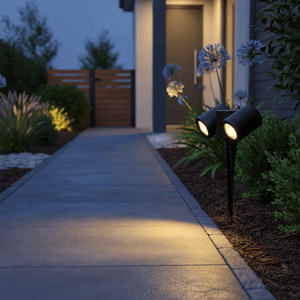 Dual-head spike light mounted in mulch, lighting a concrete pathway beside a house entrance at dusk.