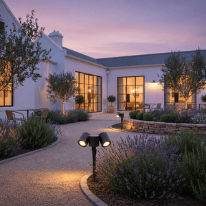Black dual‑head garden spike light lighting a gravel path beside lavender, with a white house and windows behind.