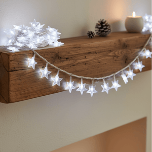 LED fairy string lights shaped like stars draped over a wooden mantel, with a candle and pinecones nearby.