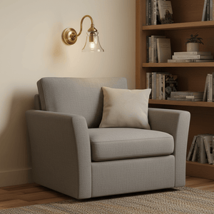 Antique brass wall lamp with clear glass diffuser above a cozy gray armchair in a warm interior setting.
