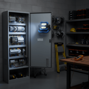 Blue and black rechargeable LED work light magnetically attached to an electrical cabinet door in a workshop.
