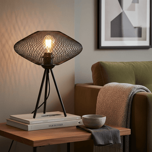 Black mesh table lamp on a wooden side table with books and a bowl, casting warm light in a cozy living room.