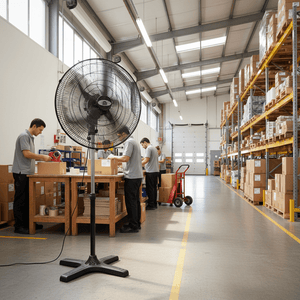 Large black industrial floor fan standing in a warehouse aisle with workers packing boxes at tables.