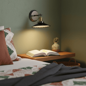 A retro industrial wall reading light mounted above a wooden bedside table with an open book and decorative items.